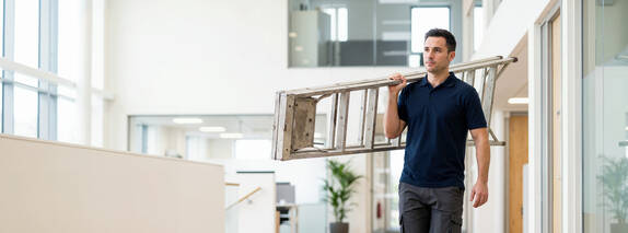 Facility Management: Person mit einer Leiter in einem Bürogebäude.