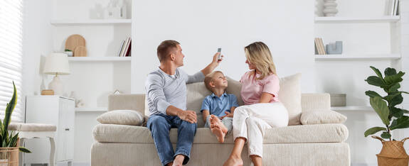 Eine Familie sitzt auf einer Couch. Der Vater bedient eine Fernbedienung.