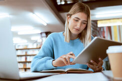 Eine junge Frau sitzt in einer Bibliothek und sieht sich etwas auf einem Tablet an