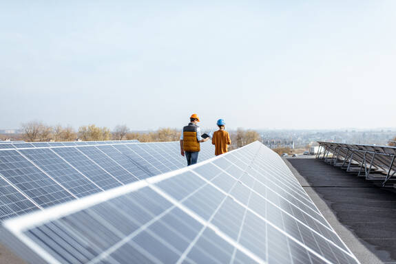 Zwei Personen mit Bauhelmen auf einem Flachdach. Sie gehen zwischen Photovoltaikmodulen.
