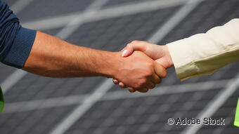 Das Bild zeigt zwei Menschen, die sich die Hand geben und im Hintergrund ist eine Photovoltaik Analge zu sehen.